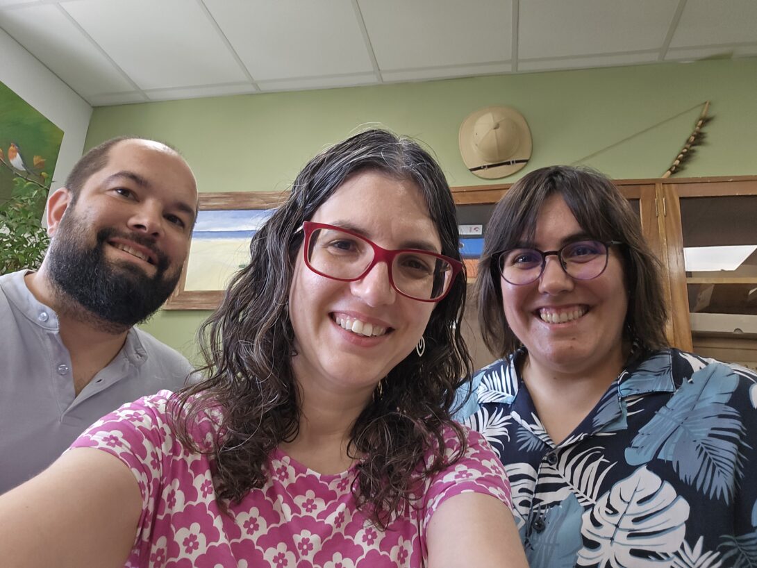 Three people smiling in a room, wearing glasses. The woman in pink is Dr. Christina Nicholas. The background includes a wall, art pieces, and a safari hat. The atmosphere is cheerful.