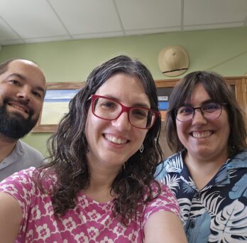 Three people smiling in a room, wearing glasses. The woman in pink is Dr. Christina Nicholas. The background includes a wall, art pieces, and a safari hat. The atmosphere is cheerful.
                  